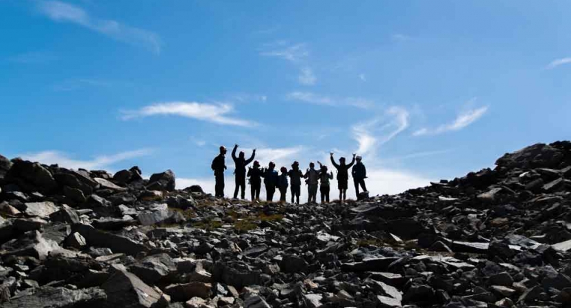 A group of students stand along a rocky ridge under blue skies. A couple of them raise their arms in the air in an apparent gesture of success. 